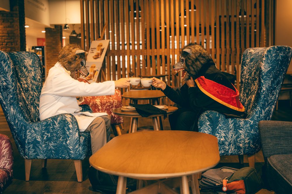 Two males dressed in a University of Wolverhampton graduation robe and lab coat. They are both wearing wolf masks. They are sat in a coffee shop in a 'cheers' motion with their coffee cups.
