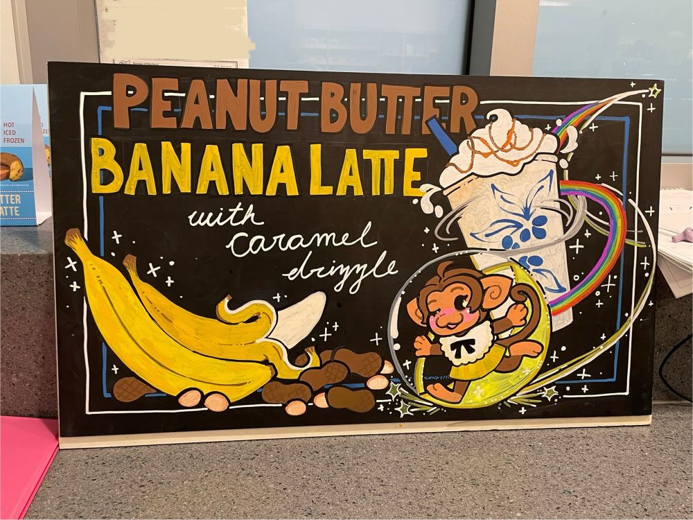 coffee shop chalk board art advertising a peanut butter and banana flavored drink, with Palette from Super Monkey Ball drawn in running around a frozen latte