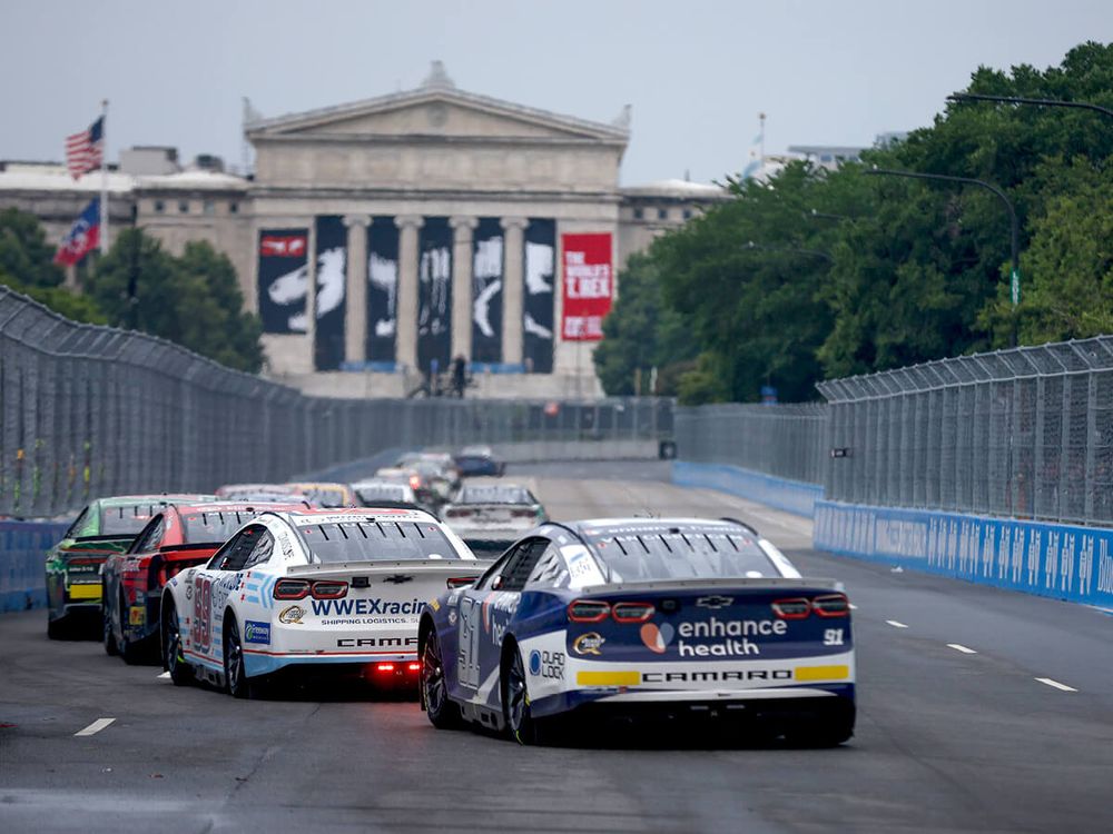 A group of NASCAR stock cars racing in front of a large building with pillars and a dinosaur in the background 
