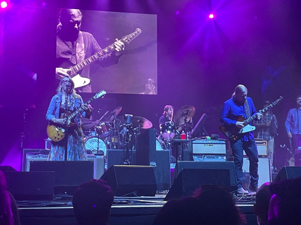 An image of Tedeschi Trucks Band, a 12(?!) person band led by Susan Tedeschi, a singer and guitarist on the stage in a floral dress and jean jacket holding a guitar, and Derek Trucks, a tall man with long blonde hair wearing a bright blue shirt holding a guitar. He is on the large screen in the background as he is playing a guitar solo. 
