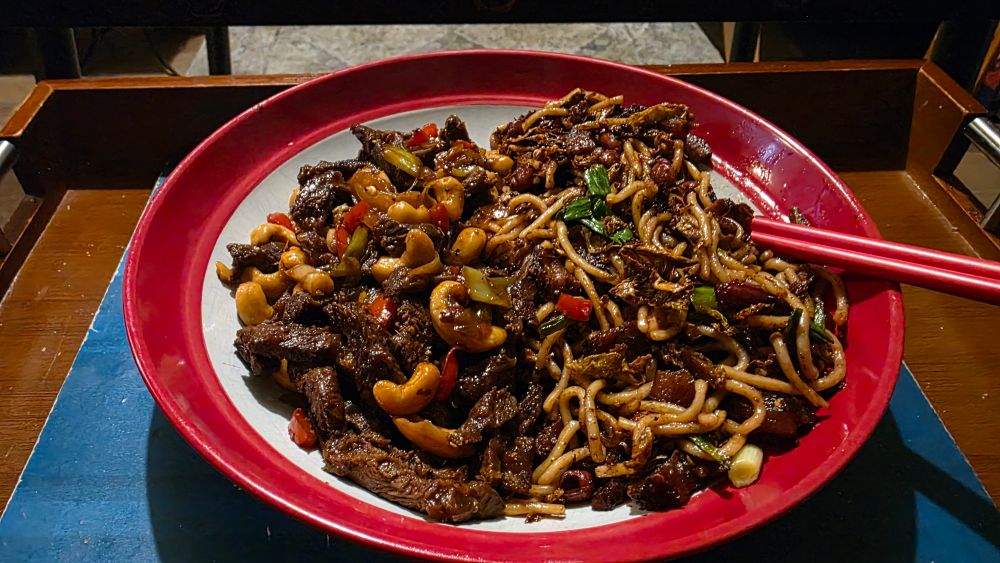 Crispy pork noodles w/ Chinese cabbage and peanuts, alongside beef and peppers w/ ginger and cashew, sitting in a red rimmed flat bowl, with red ceramic chopsticks to the right. The bowl sits on a blue place mat atop a cherry wood dinner tray with inset metal handles in a Japanese style