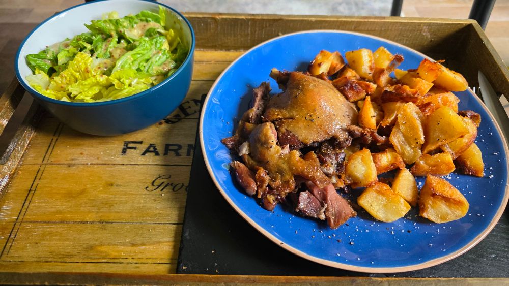 A wooden farmer's market tray on which sits a large slate mat in the bottom right corner. Atop that is an azure blue plate with crispy confit'd duck leg and a mound of golden duck fat roasted potatoes liberally sprinkled with sel de Guérande. Top left corner of the tray sits a deep teal bowl of romaine lettuce dressed with a honey, lemon and wholegrain mustard sauce. Out of shot is a large glass of 2018 Morgon, but trust me, it's there 