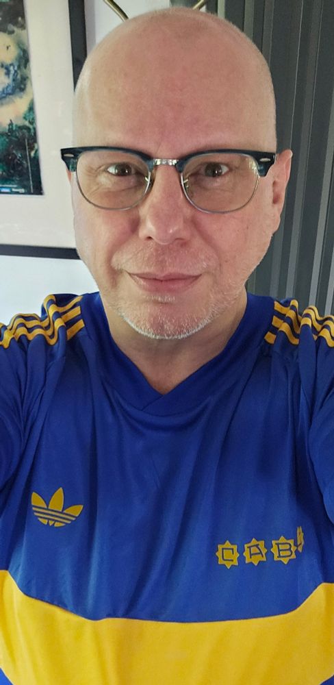 Same bald man, same glasses, different shirt (plot twist!)

Royal blue with horizontal yellow stripe, its the Boca Juniors shirt as worn the legendary 1981-82 season by God*

*Diego Maradona 