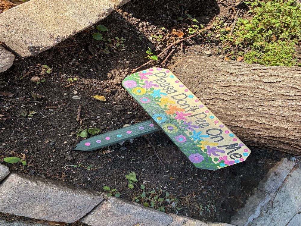 A hand painted sign laying in dirt that reads “Please don’t pee on me.”
