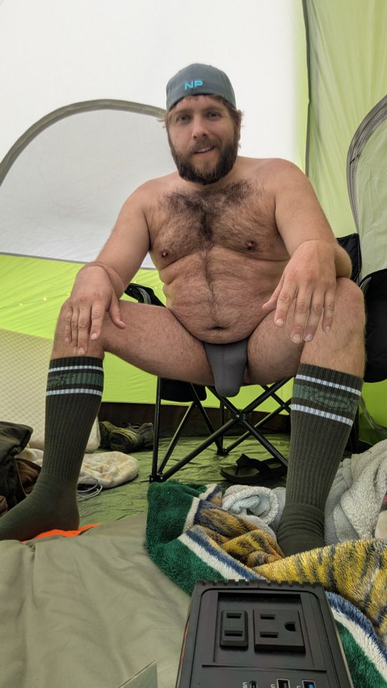 Man only in a backwards hat, jock, and socks. Sitting inside a tent.
