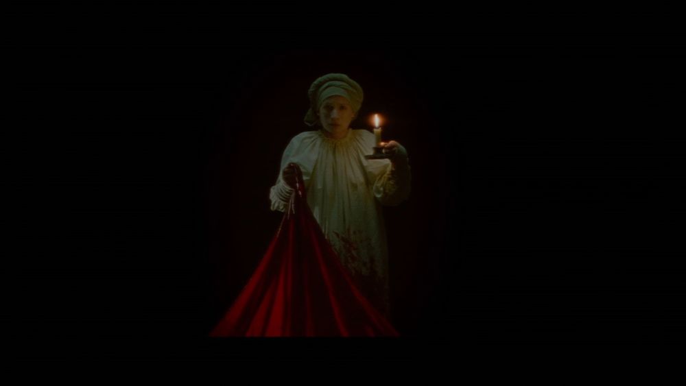 Screenshot from the movie "The Vourdalak" showing a man in pajamas holding a candle and a red velvet sheet in a pitch black room.