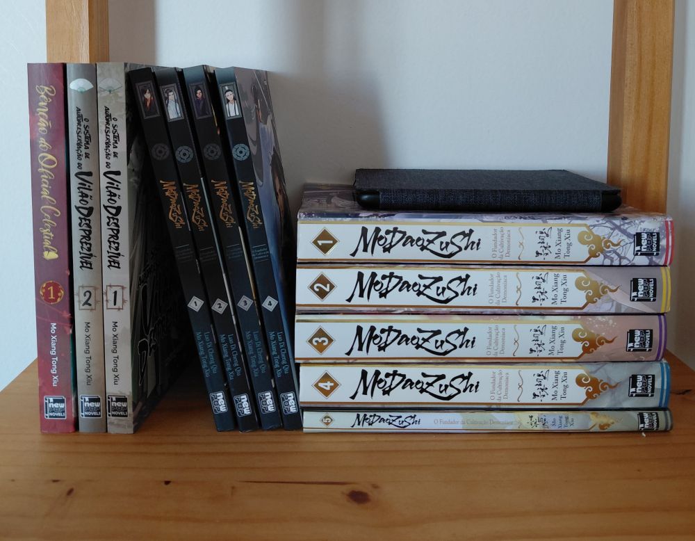 Prateleira de livros com as versões brasileiras das novels MDZS, TGCF e SVSSS, da MXTX. 