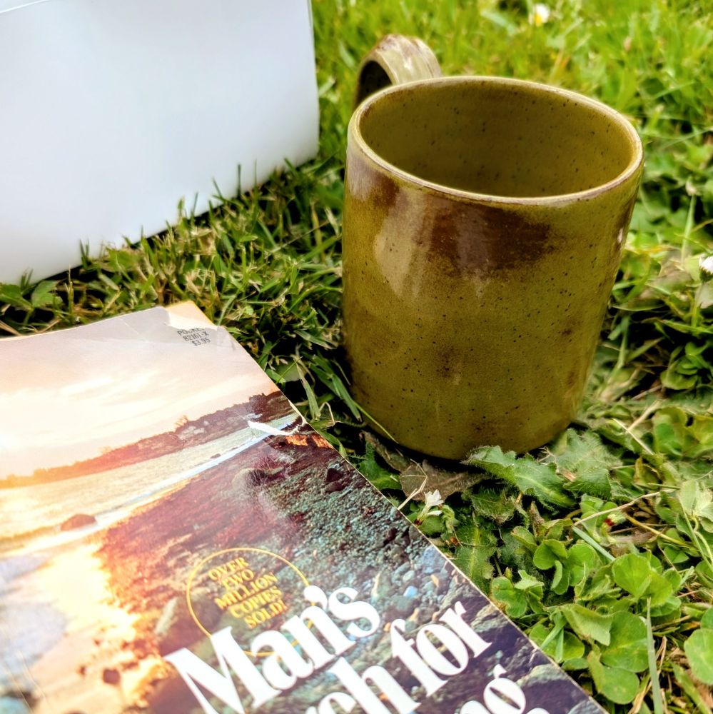 A hand-thrown mug in a dark olive green sits on bright green grass. A book lies splayed open with the cover showing... because I'm a monster and I crack spines. In the upper left corner is a mysterious block of white. It's a treat box that used to contain a homemade cinnamon bun and a rhubarb fritter...but I ate those so nobody would drool over their deliciousness in a picture. You're welcome.