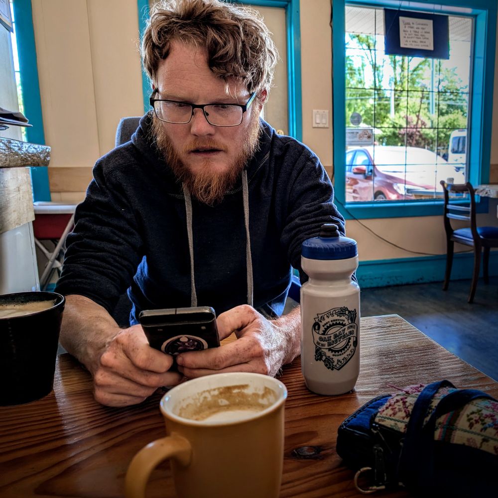 A scruffy redheaded man with a swoop of hair over one side of his forehead stairs intently at his phone in a quaint little coffee shop. Beside him is his black mug of coffee and a blue and gray water bottle.  In the foreground are a yellow mug of coffee and a small embroidered purse. Behind him is a window framed in bright teal against butter yellow walls.