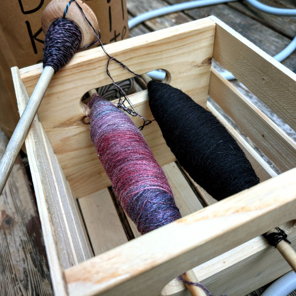 Two spools of single-ply hand-spun yarn are wound thickly onto two long wooden dowels. The spool on the left is variegated purples, blues, and a deep wine red. The spool on the right is black. The ends of each spool sit neatly in the hand-holes of a small wooden crate...a sort of make-shift lazy kate if you will. Lying across the upper left corner of the crate is a handmade wooden drop spindle with the beginnings of two-ply stripey yarn.