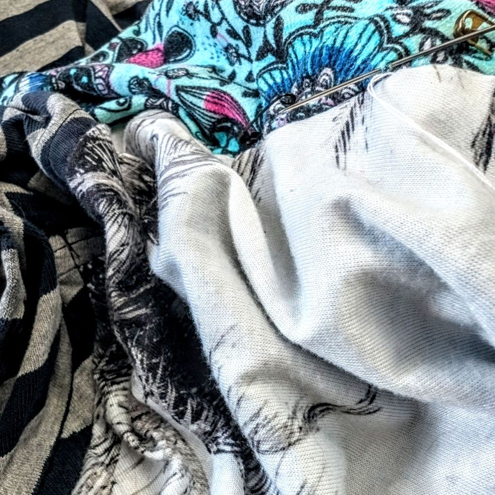 A crumpled pile of fabrics...we have grey and black stripes along the left edge, a teal fabric with pink and blue highlights and black flower doodles across the top, and the middle and bottom right is taken up by a white fabric with black and grey palm leaves that kinda look like feathers. A needle and brass safety pin stick out of the teal fabric near the top.