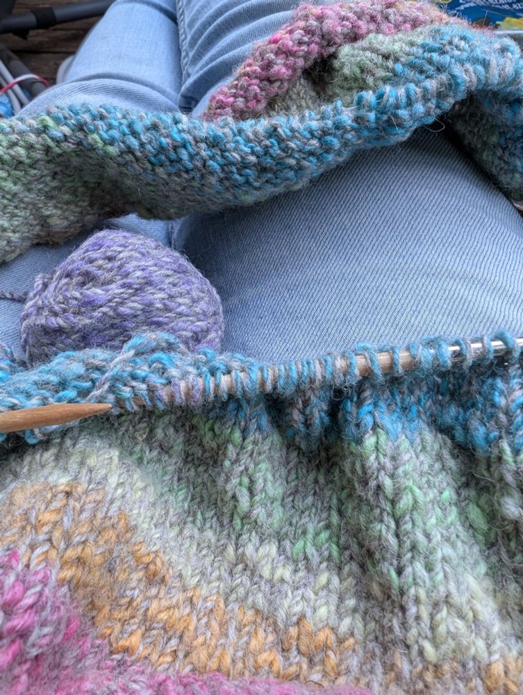 Knitted fabric in soft rainbow stripes laying across my legs. The stitches are uneven because this was the first ball of yarn I've ever spun, so it has lots of lumpy character bits. I plied it with grey wool, too, so that adds more visual texture.