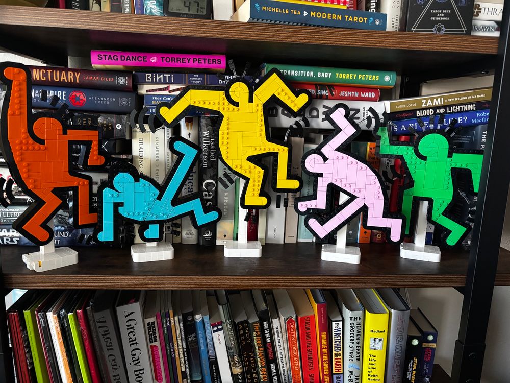 5 Lego figurines in the shape of Keith Haring’s Dancing Figurines. From left to right they are red/orange, teal blue, yellow, light pink, and green. They held up with white stands on my bookshelf in front of TOO MANY books to name. 