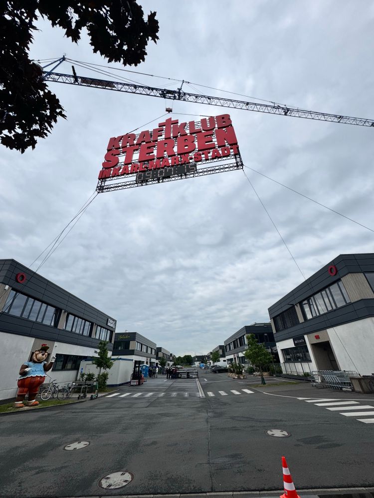 Ein Schild an einem Kran: "KRAFTKLUB – STERBEN IN KARL-MARX-STADT" über eine noch leeren Bühne im Segro Gewerbepark Bickendorf, wo die Band heute, Donnerstag 5.6.2025 um 19 Uhr, alle zu einem kostenlosen LIVE-KONZERT einlädt! 