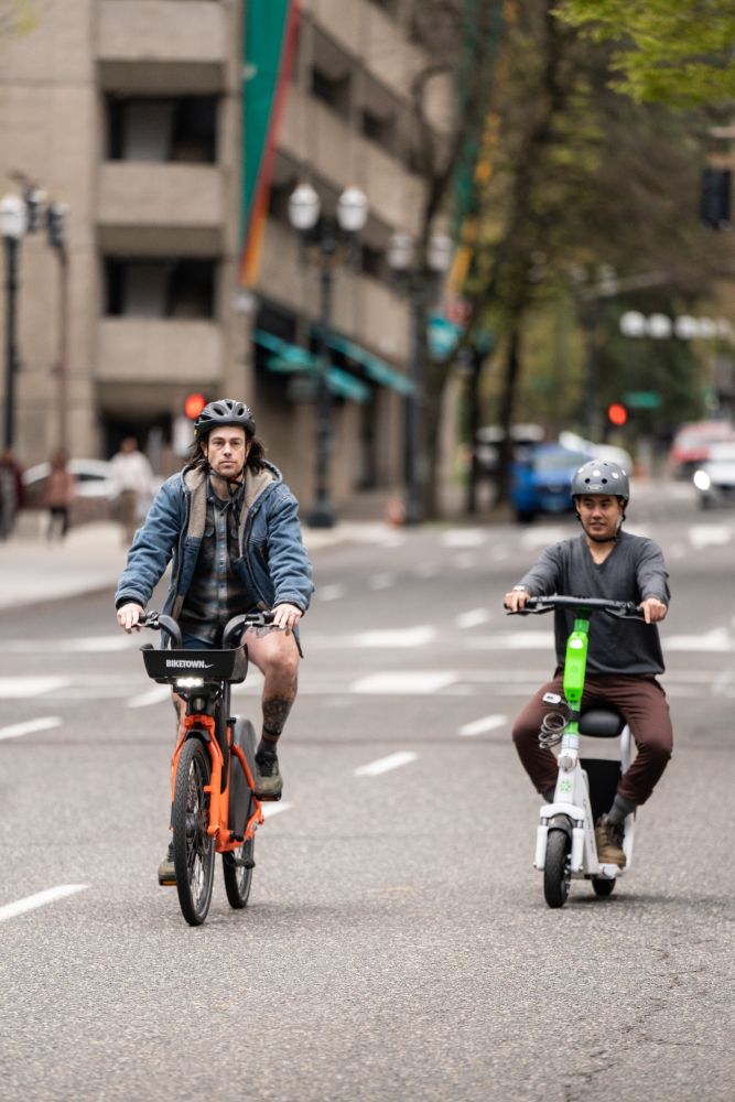 One person rides a BIKETOWN bike while another person rides a Lime e-scooter in downtown Portland. 