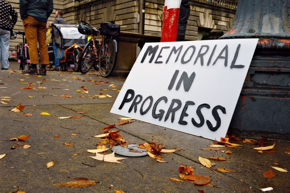 A white sign leaning against the base of a light pole with painted black text reads, “memorial in progress.” 