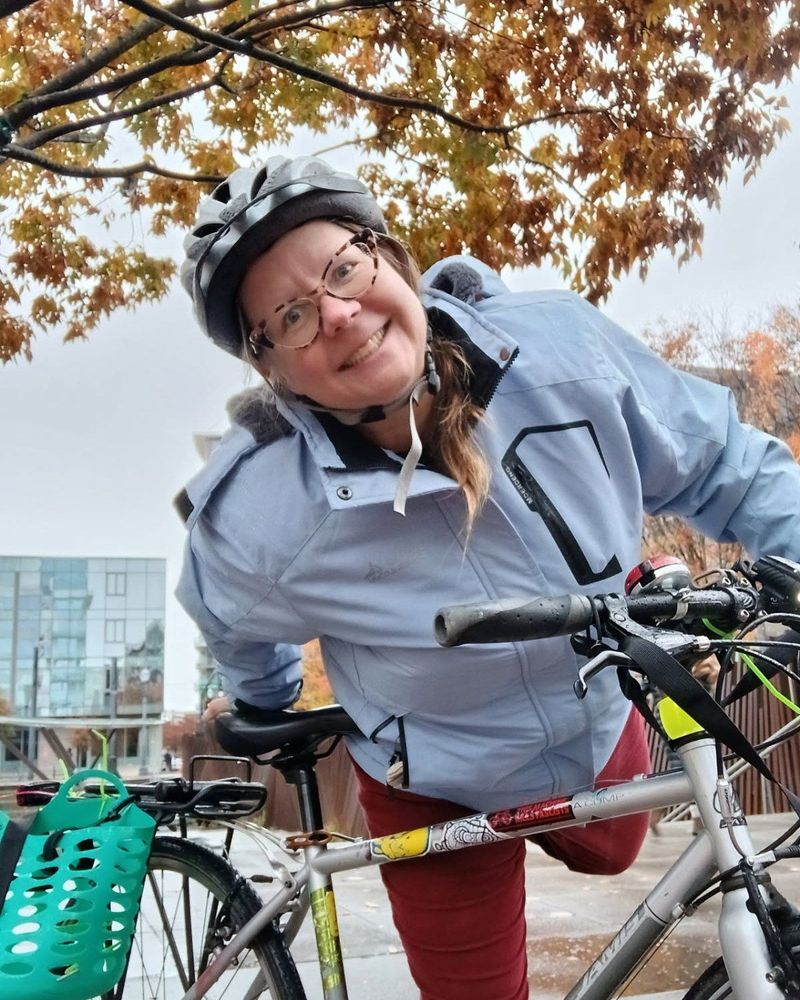 A person wears a grey raincoat and stands with their bike under a colorful, fall tree. 