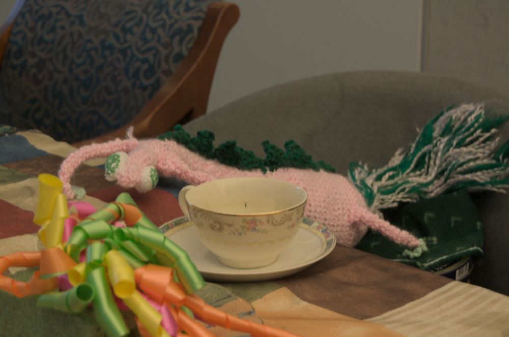 Blorplesnorf - a pink creature with green back spines, six legs, and two bulbous eyes sits in front of a delicate floral-print tea cup and saucer.