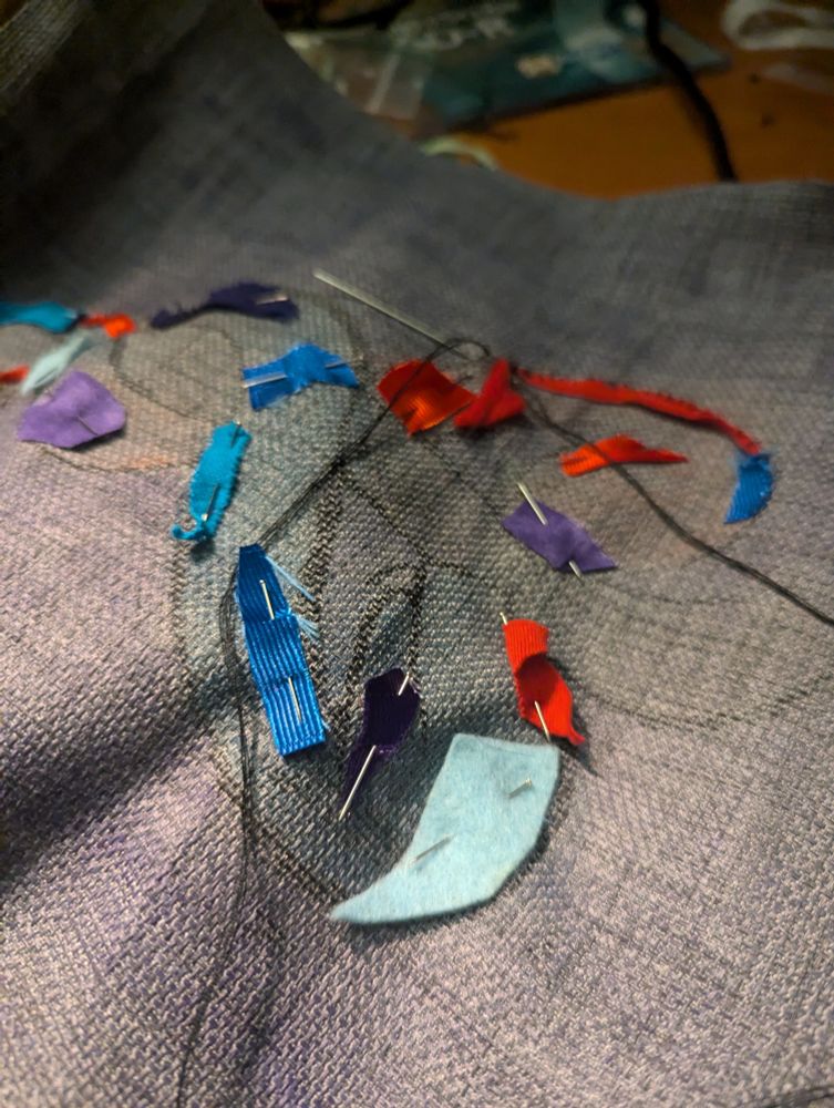 The colorful snake design from the first image, now with scraps of red, blue, and purple cloth pinned to the snakes' skin. A needle and thread sits off to the side, next to two pieces that have been sewed down along the contours of the red snake's head. 