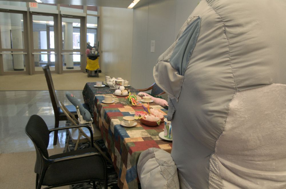 View of the table over Wesley's shoulders. Shows the long table, with patchwork table clothes and platters of weird food and the set of chairs. Some are empty, some are occupied by strange creatures.