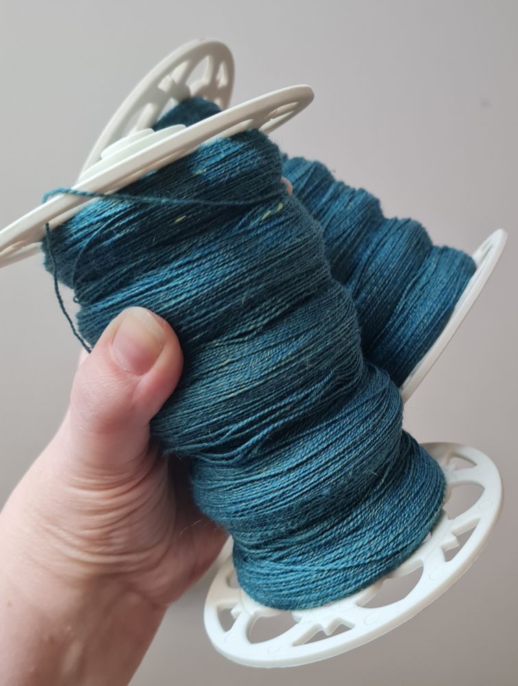 A hand holding two white spinning wheel bobbins, each bobbin is filled with handspun teal blue two ply laceweight yarn.