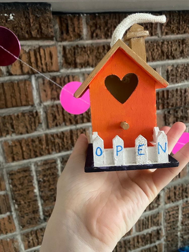 Front of birdhouse with orange painted door and white picket fence with the letters “O P E N” written on the panels