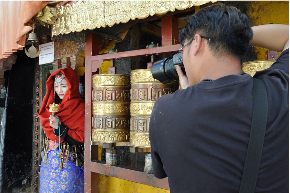 A tourist dressed in a Tibetan costume poses by copper prayer wheels as a  photographer  aims his camera lens at her.