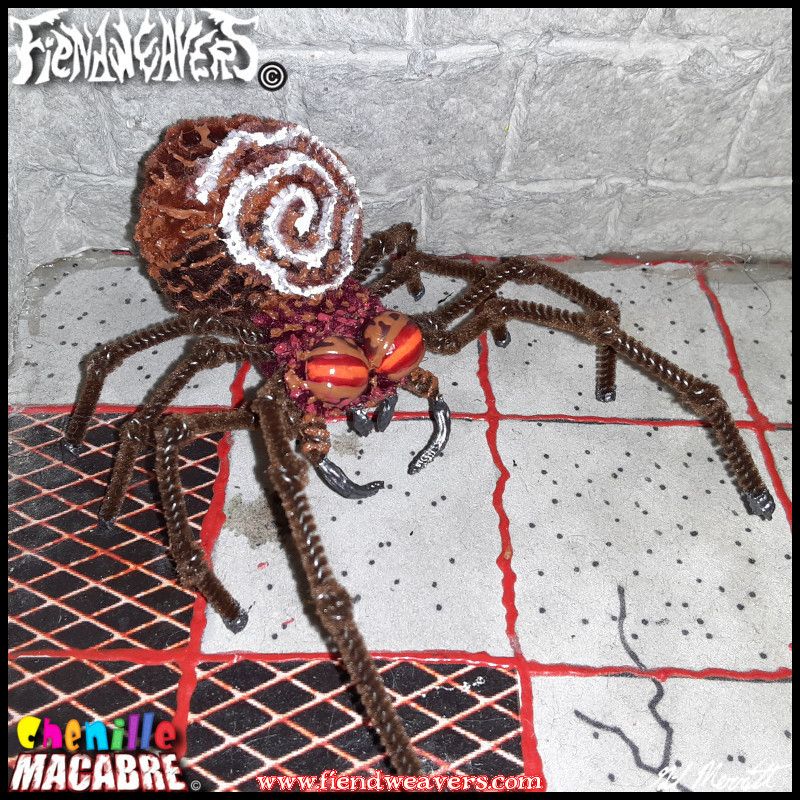 A pipe cleaner figure I built of a spider I call the Spiral Deathtrap. She has a brown abdomen and legs, a burgundy head, large piercing red eyes, and a white spiral pattern in her abdomen. Her forelegs are much longer than her hind legs. She stands 2" (5.08 cm) high with a 4" (10.16 cm) legspan.