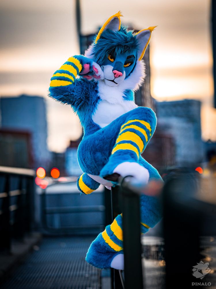 A feminine lynx fursuiter sits on a metallic railing. One leg is resting on the railing, while the other is on the side. She has her handpaw near her head in a confident pose and looks directly at the camera. In the background, there are large concrete buildings.