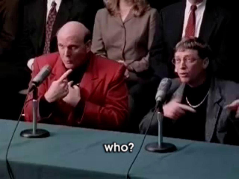 a man in a red jacket is sitting at a table talking into a microphone and asking who .