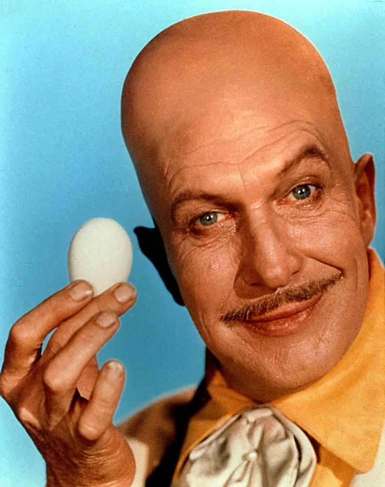 vincent price as the villain known as egghead from the batman 66 tv series. he has a bald, egg shaped head, though less egg shaped than marc andreessen's head oddly enough, and a moustache and is wearing a yellow top while holding an egg for the viewer like it is a precious gem or something he is about to do a magic trick with.