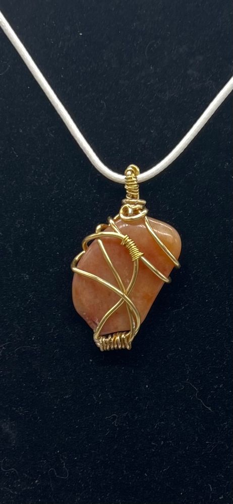Closeup of coral colored tumbled stone pendant, wrapped with gold-toned wire and strung on a white leather cord. 
