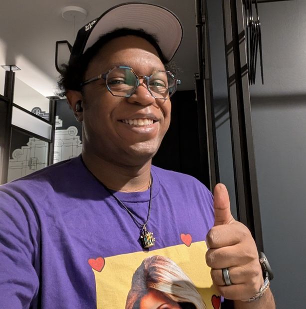 Sparrow, smiling with a purple Topanga shirt, a black baseball cap, and the birdcage necklace. 