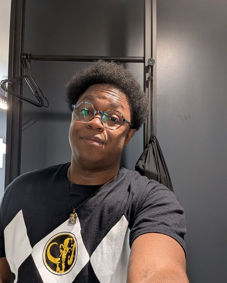 Sparrow, smiling wearing a black power ranger t-shirt, heptagonal glasses, and a bird cage necklace. 