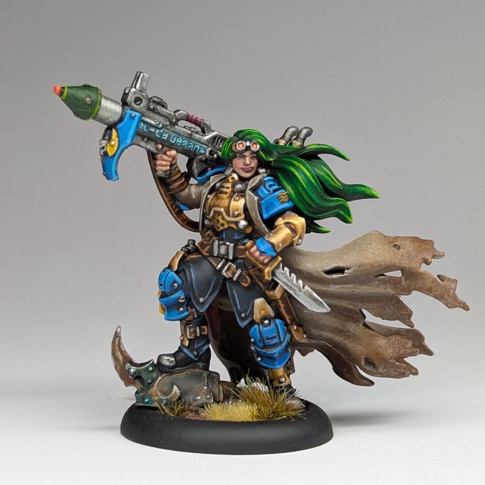 A painted miniature of a warmachine warcaster. She has green hair, and wears blue and gold armour with a long tattered brown trenchcoat. She wields an arcane rocket launcher