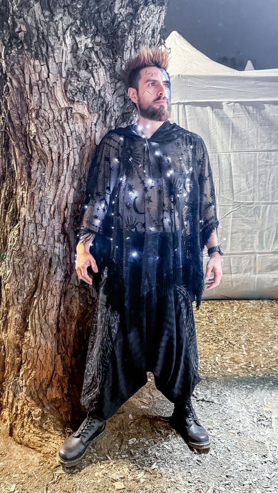 Dan surge como um oráculo noturno no Medieval Experience, encostado em um grande tronco de árvore iluminado pela luz branca que destaca sua figura. Ele está com o cabelo ruivo espetado para cima, barba cheia igualmente ruiva e o rosto marcado com pinturas em azul escuro e traços que remetem a constelações, reforçando a aura mística da caracterização. Veste um poncho preto de renda transparente estampado com luas, estrelas e símbolos celestes, decorado com pequenas luzes que brilham como pontos estelares sobre o tecido. A peça tem franjas soltas que caem sobre as calças largas, também pretas, de estilo saruel. Nos pés, botas pretas pesadas completam o visual. O braço esquerdo exibe um relógio e pulseiras discretas, enquanto o corpo inteiro assume uma postura firme, de quem incorpora o personagem com intensidade. Ao fundo, além da árvore, há uma tenda branca de tecido iluminada, reforçando o clima de feira medieval noturna. O conjunto da cena mistura fantasia e mistério, com Dan transformado em uma figura enigmática e quase sobrenatural.