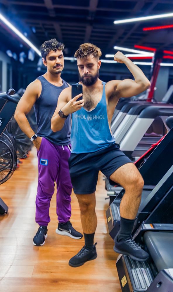 Rodrigo e Dan aparecem lado a lado na academia, logo após um treino intenso de bíceps. À esquerda, Rodrigo exibe braços definidos e ombros largos, vestindo uma regata preta que realça seu peitoral marcado e seu visual enxuto. Seu rosto sério contrasta com os cachos escuros bem definidos e o bigode fino com cavanhaque. Ele usa uma calça roxa estilosa com detalhe azul e tênis pretos com sola branca.

Ao lado, Dan assume uma pose mais performática: com o joelho apoiado na lateral da esteira, exibe o braço direito flexionado, destacando o volume do bíceps e o antebraço espesso. Sua regata azul extremamente cavada expõe grande parte do peitoral e da lateral do tronco, deixando à mostra a pele clara, os pelos densos e o contorno do músculo peitoral. A regata solta permite ver até o início da barriga, enquanto o short preto e as meias altas pretas emolduram suas pernas grossas, cobertas por pelos visíveis e bem distribuídos. Seu rosto está sério e concentrado, com barba cheia e cabelo castanho claro levemente bagunçado, voltado para o espelho enquanto segura o celular. A iluminação da academia realça os contornos físicos e dá ao cenário um ar de intensidade e foco pós-treino.