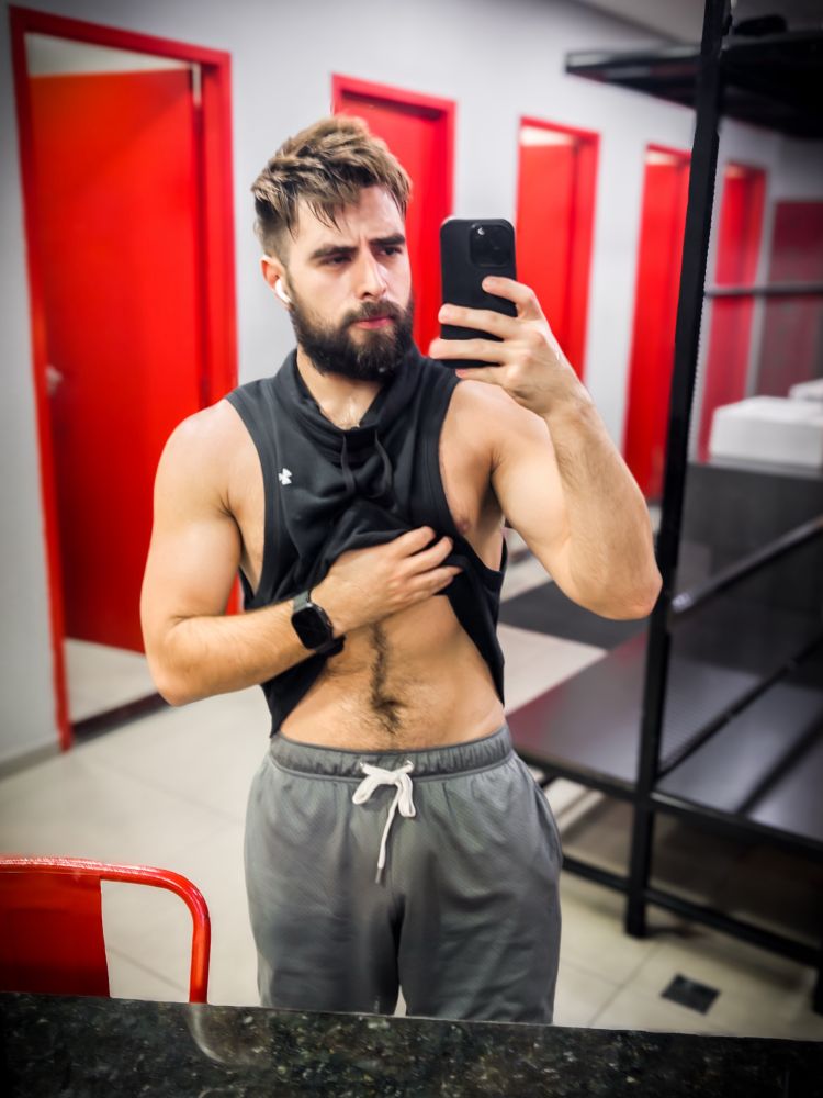 Dan surge em frente ao espelho de um vestiário, levantando a própria regata preta cavada para exibir parte do abdômen. A regata justa ressalta seus ombros largos e os braços volumosos, com veias discretas que acompanham o contorno dos bíceps. O peitoral parcialmente exposto é coberto por uma camada de pelos escuros, que se estendem até a barriga e descem em uma faixa central definida. A região do abdômen, embora parcialmente coberta, revela a textura firme da pele e a presença de músculos levemente definidos sob uma camada discreta de pelos. Dan usa uma bermuda cinza, amarrada por cordões brancos. O rosto exibe expressão concentrada, as sobrancelhas franzidas e lábios cerrados, emoldurado por uma barba espessa. Os cabelos castanhos claros estão bagunçados, com volume e movimento, reforçando o ar naturalmente intenso da cena.