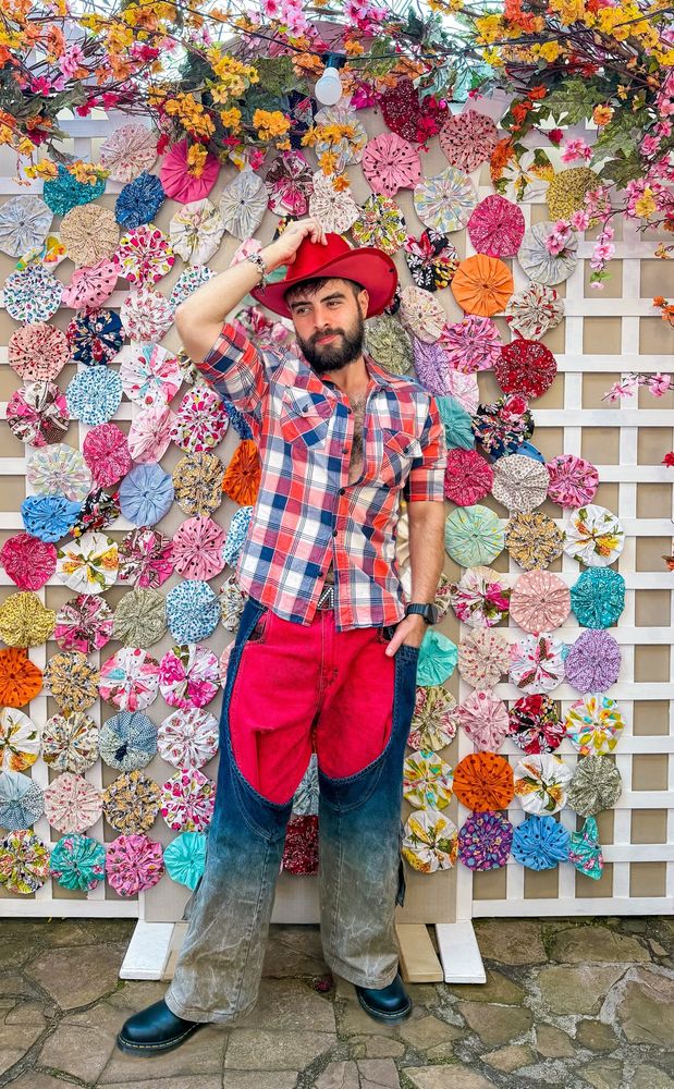 Dan domina a cena com sua postura confiante e leve sorriso, combinando carisma e irreverência com naturalidade. Em destaque, ele posa com a cabeça levemente inclinada e um sorriso contido nos lábios, segurando a aba de um chapéu vermelho de vaqueiro com a mão direita, num gesto descontraído que acentua seu ar brincalhão. Seu rosto é emoldurado por uma barba cheia e bem cuidada, e os cabelos castanho-claros estão levemente bagunçados, dando um charme casual à sua aparência.
Com o tórax parcialmente exposto pela camisa xadrez em tons azuis, vermelhoz e brancos, que está entreaberta, Dan revela parte de seus pelos peitorais, além dos ombros largos e braços definidos que se destacam com as mangas dobradas. O visual evidencia sua forma, marcada por sua presença magnética.
A bermuda jeans vermelha, de modelagem solta, é o centro do figurino e cobre até pouco acima dos joelhos. Sobre ela, Dan usa perneiras jeans azul-escuras que descem soltas pelas laterais das pernas e vão desbotando até a barra, criando um efeito visual rústico e estilizado. As botas pretas reforçam o aspecto tradicional do look com firmeza.
Ao fundo, um painel branco em treliça serve como suporte para uma profusão de fuxicos coloridos de tecido, formando uma moldura vibrante de flores artesanais e galhos floridos, em tons de rosa, amarelo, laranja e lilás. Mas, mesmo diante desse cenário festivo e carregado de detalhes, é Dan quem atrai todos os olhares, com seu magnetismo e presença marcante.