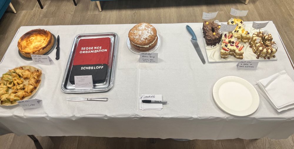 Cakes on a table including a baked cheesecake, pastries, a Schegloff book shaped cake, Victoria Sponge, and cakes in the shape of the letters in DARG. 