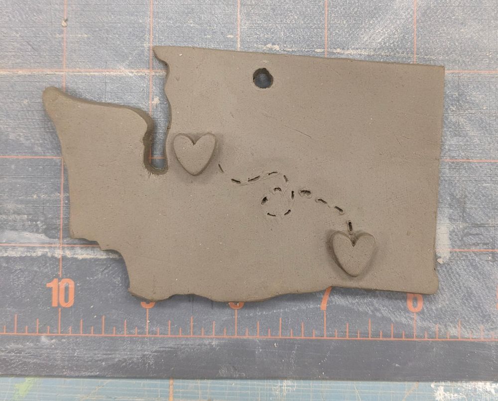 An unfired ceramic ornament that is in the shape of Washington State with 2 heart shapes symbolizing 2 locations with a dashed line between them symbolizing moving.