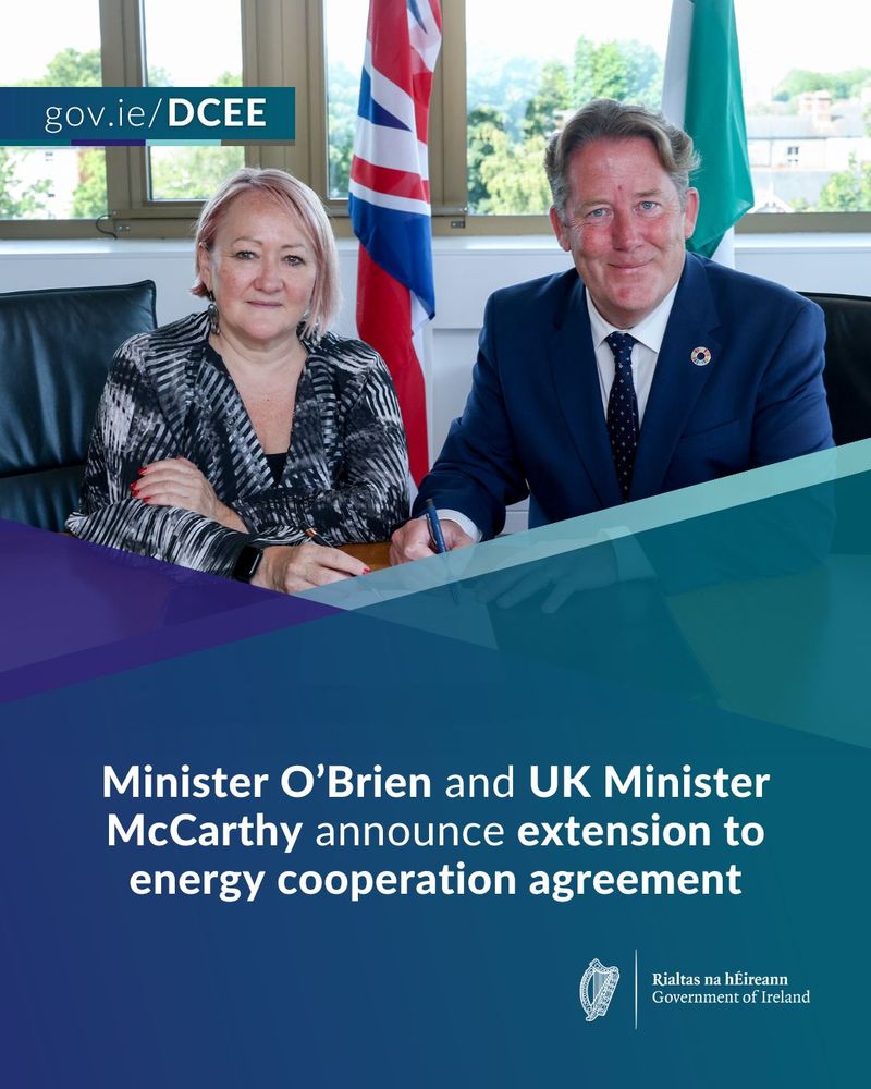 Two government ministers sit at a table signing an energy cooperation agreement, with flags of the UK and Ireland visible in the background.