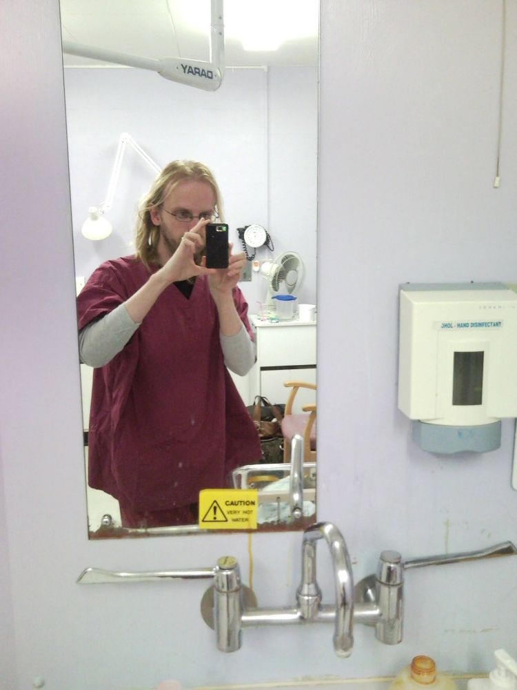 Me, at the time a skinny white man with shoulder length blond hair, gingery beard and glasses. I'm taking a mirror selfie in a hospital room, I'm wearing burgundy scrubs. 