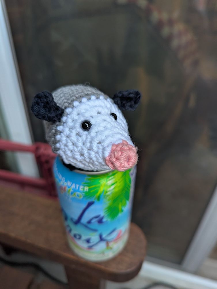 Amigurumi possum on top of a blue can of fizzy water. Front view of critter. 