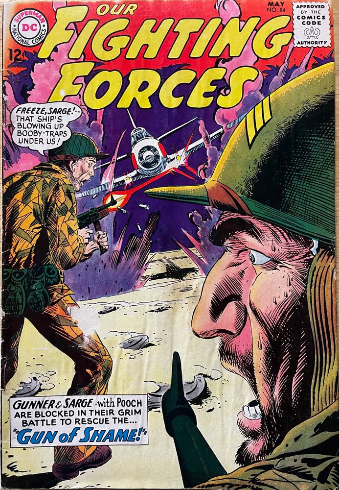 Two American soldiers are standing in a field as an airplane is flying toward them while firing its guns. 