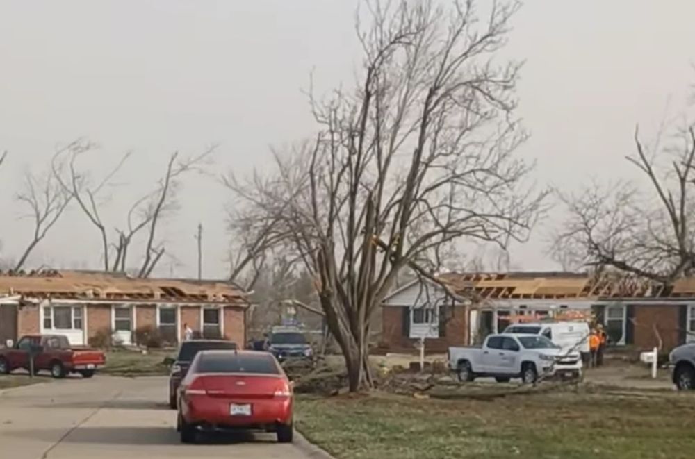 Picture of two houses in the distance with their roofs blown off by a tornado. The trees in the area have been damaged as well. 