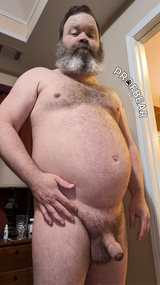 A naked, bearded CIS man standing in a bedroom.