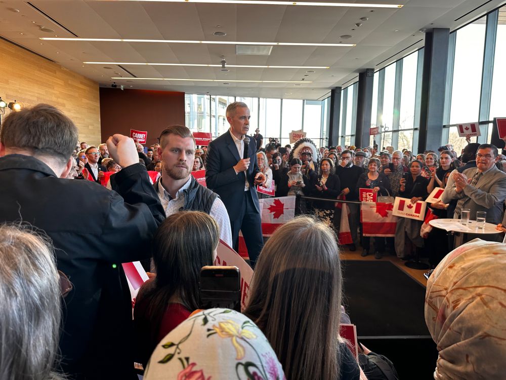 Prime Minister Mark Kearney speaks at a packed campaign event in Saskatoon. Enthusiastic supporters of all ages and ethnicities, hold signs and support.
