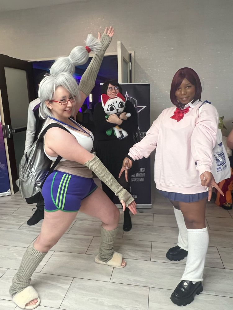 Seiko okarun and momo cosplayers. Seiko is showing off momo and momo is doing double peace signs while okarun and turbo granny hide in the back 