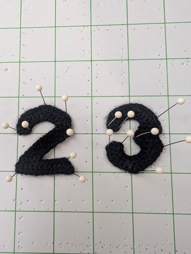 A number 2, and a number 3 crocheted in black yarn are pinned to a blocking board as they dry from blocking. 